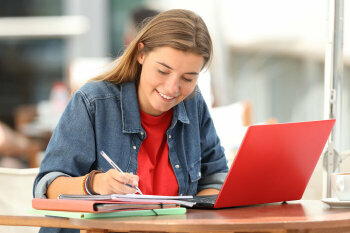 Ein lächelndes Mädchen sitzt vor ihrem aufgeklappten Laptop und macht sich Notizen.
